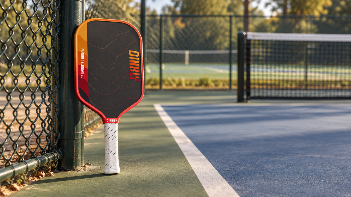 Dinkly Fusion Elongated paddle standing beside a pickleball court fence